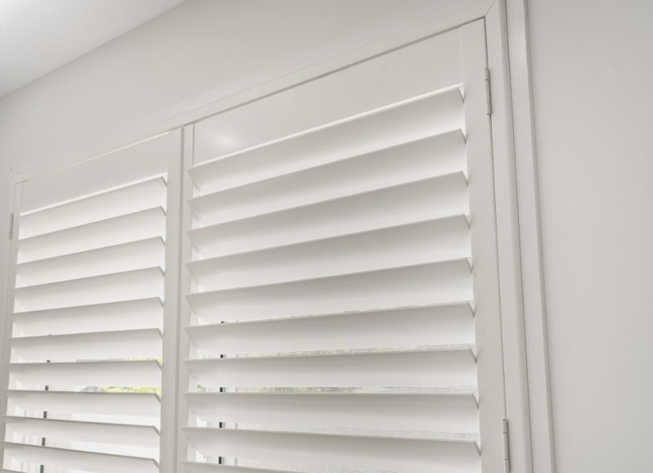 Indoor PVC shutters partially open on a sunny day, allowing light to filter through, creating a soft ambiance in the room. The clean, modern design complements the neutral wall color, adding an elegant touch to the space.