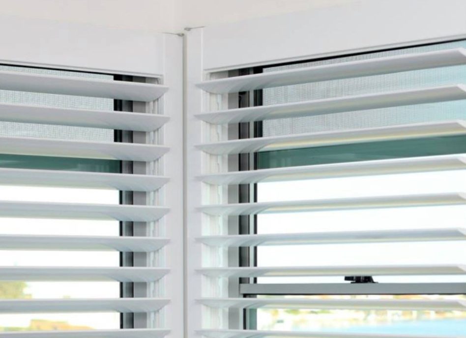 Close-up of indoor PVC shutters partially open on two adjacent windows. The background view outside is blurred but reveals hints of blue sky and greenery. These shutters gracefully allow natural light to filter into the room.