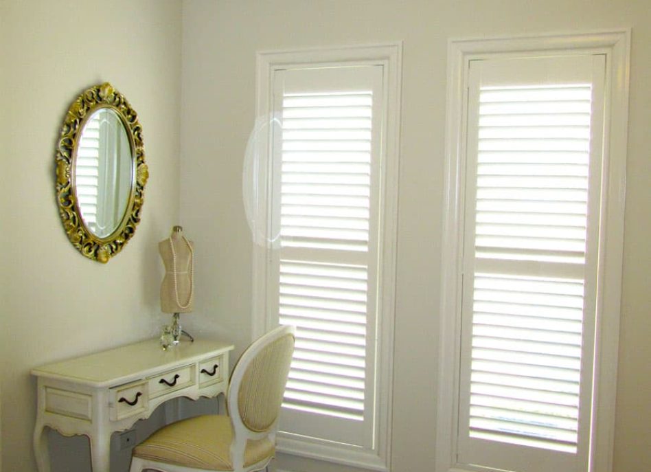 A bedroom corner with two windows featuring white shutters, a white vanity with a mirror, a chair, and a chandelier above. Part of a bed with pink and white bedding is visible.