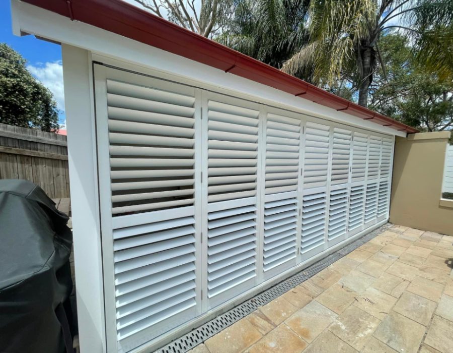 A white louvered shutter door made of durable outdoor aluminum is installed on a patio with a red roof. Palm trees and a wooden fence frame the background, while a barbecue grill peeks in from the left. The patio floor is elegantly paved with tiles.