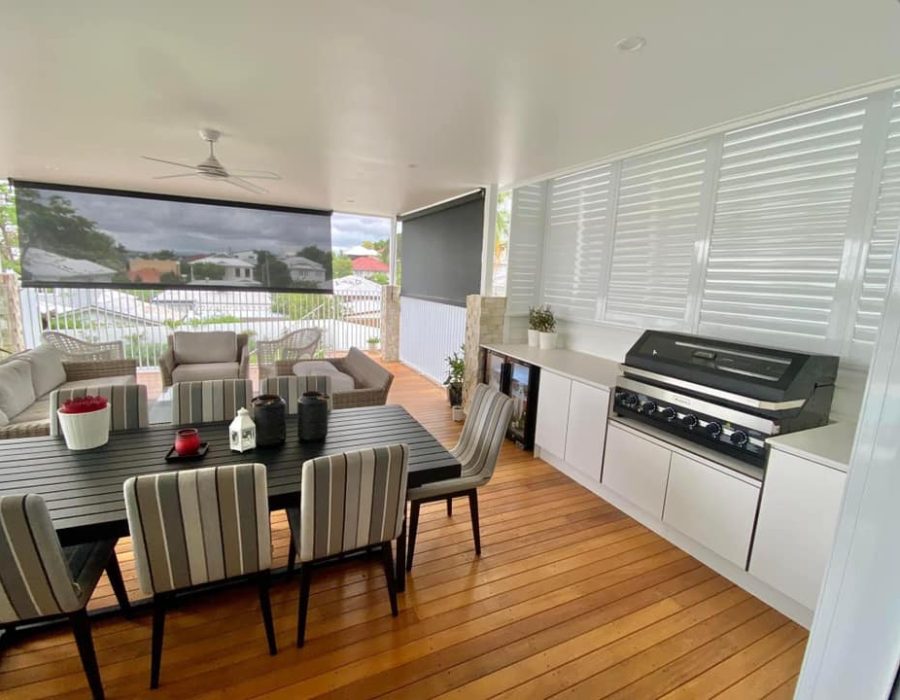 A spacious, covered outdoor patio features a wooden floor, striped cushioned chairs around a black dining table, a sofa set, and a built-in barbecue grill. Large windows and outdoor aluminum shutters allow natural light while offering a view of trees and neighboring houses.