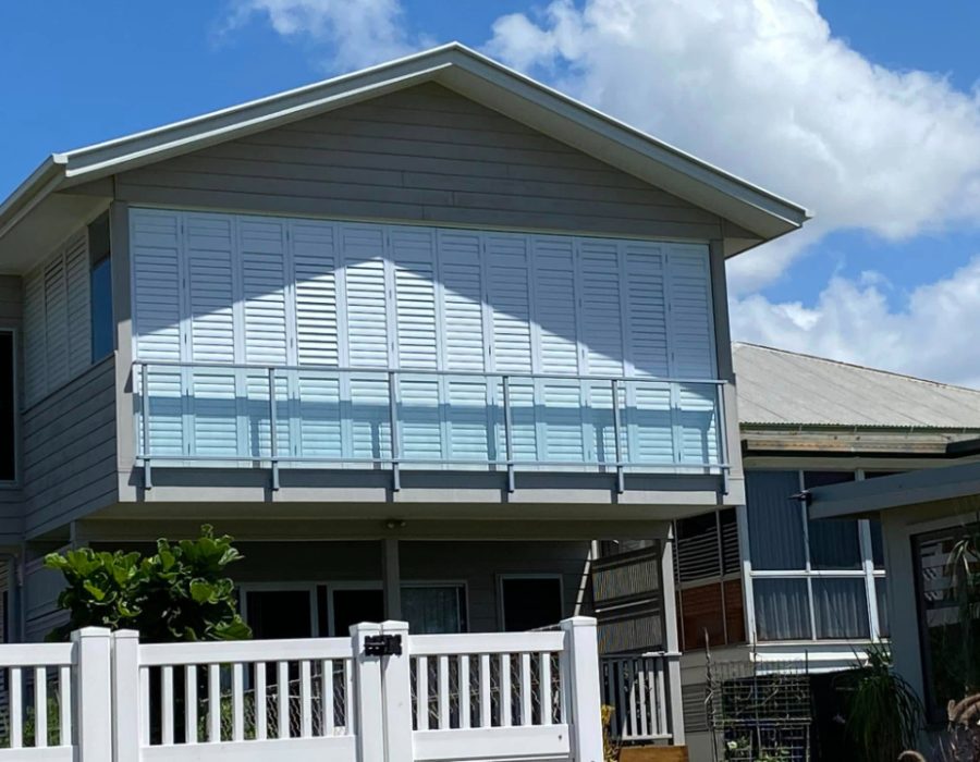 Modern two-story house with gray siding and a spacious balcony featuring sleek outdoor aluminium shutters. The front yard is enclosed by a white fence, completing the elegant look. The sky is clear with a few clouds.