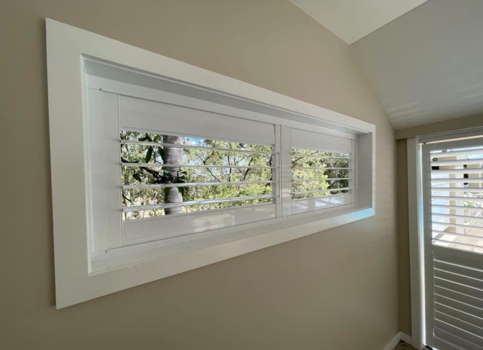 A horizontal window with white plantation shutters, complemented by outdoor aluminium shutters, partially open to reveal green trees outside. The white frame stands against beige walls, and natural light filters through the slats, illuminating the interior space.