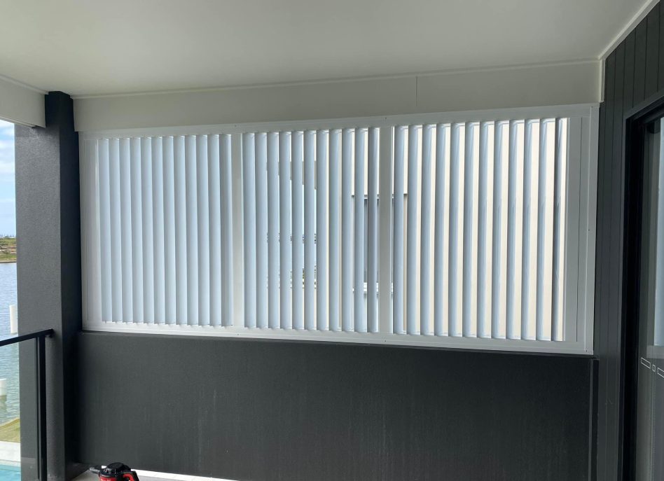 A balcony with white vertical blinds partially open, allowing light to filter through, complements the sleek outdoor aluminium shutters. The walls are dark grey, and there is a glass railing on one side. A black bag rests on the floor in the corner.