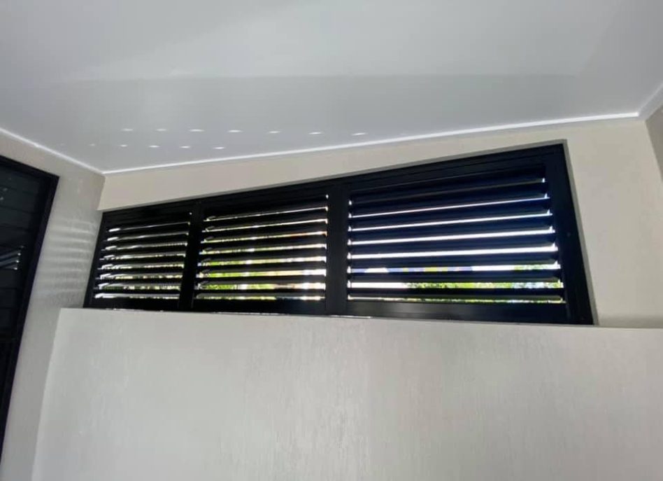 A room features high, narrow black louvers near the ceiling, reminiscent of outdoor aluminium shutters, letting in light while maintaining privacy. The walls are light-colored and the ceiling is a crisp white.