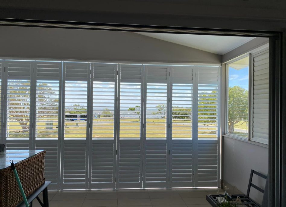 A sunlit room with white outdoor aluminium shutters covering large windows offers a view of a grassy area with trees in the distance. A wicker basket rests on the left, while a small table with items sits on the right.