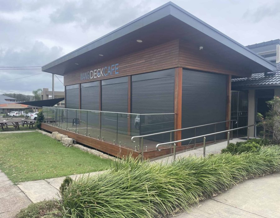 Wangi-Cafe-1–7-3-25 A modern café building with closed metal shutters, wooden exterior, glass railing, and a wheelchair-accessible ramp, surrounded by greenery under a cloudy sky.