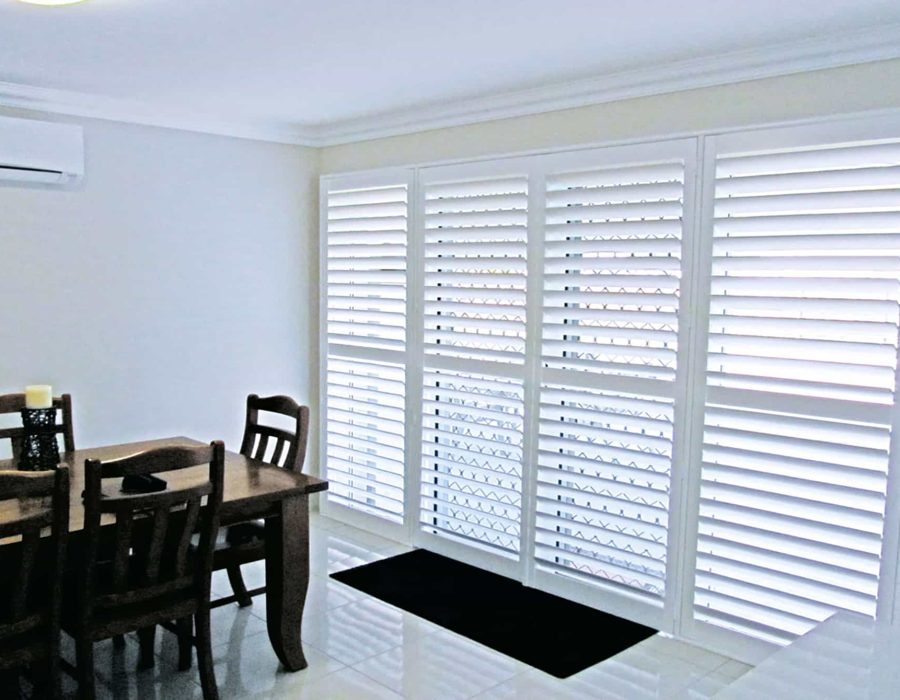 A bright dining area featuring a wooden table with six chairs and white plantation shutters covering the windows and doors. An air conditioning unit is mounted on the wall.