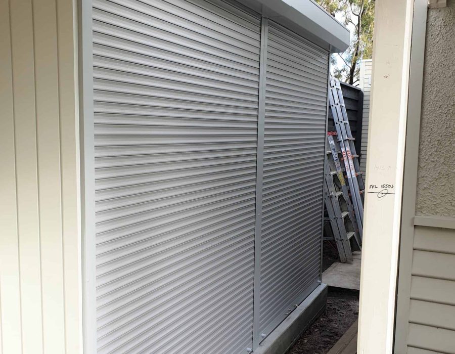 spring-operated-with-lock—1- A small outdoor shed with closed metal roller shutters, next to a house. A ladder and some construction materials are visible nearby.
